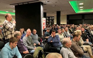 Een jongeman met een rode baard staat in een zaal vol mensen met een microfoon in zijn handen.