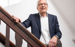 Man met grijs haar en ronde bril kijkt glimlachend in de camera. Hij staat op een trap.