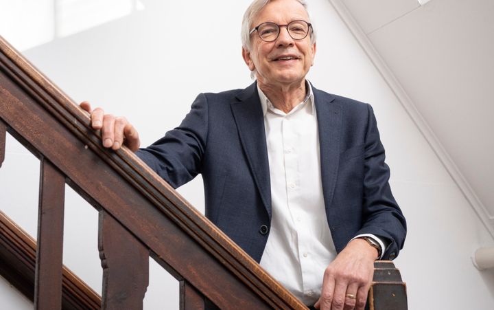 Man met grijs haar en ronde bril kijkt glimlachend in de camera. Hij staat op een trap.