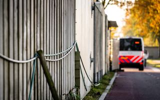 Een weg met daarlangs een houten schutting en een stenen muren, met verderop een geparkeerde witte politiebus met blauwe en feloranje strepen.