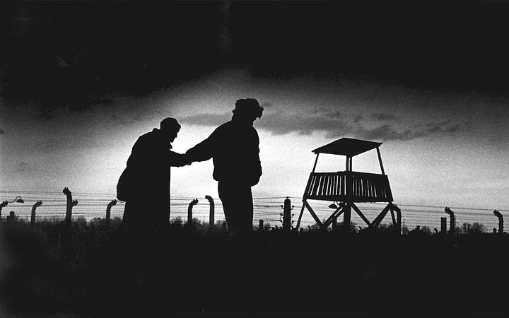 Twee donkere schimmen, hand in hand, lopen langs wachttoren en prikkeldraadversperring.