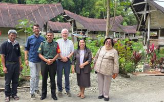 Vier mannen en twee vrouwen staan naast elkaar op een oneffen pad voor een tuin. Daarachter zijn enkele overkappingen zichtbaar.