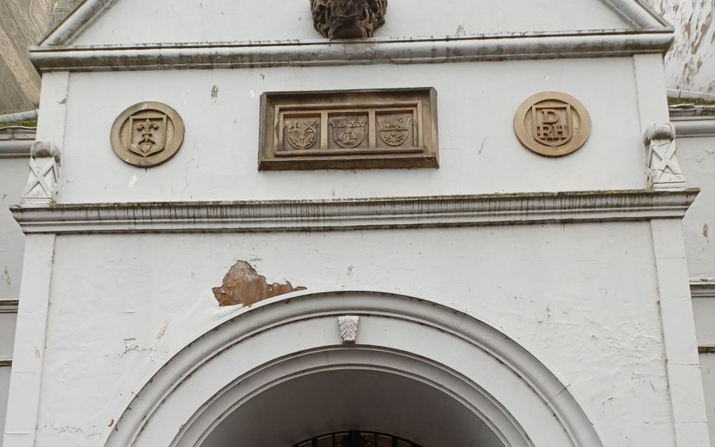 Open doorgang in een wit gebouw. Een ijzeren toegangsdeur staat half open. Boven de ijzeren deur staan woorden.