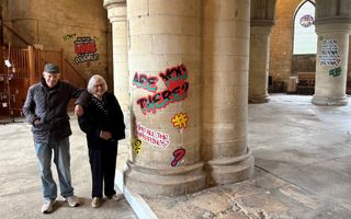 Een oudere man en vrouw bij een van de pilaren waarop in graffiti spreuken zijn aangebracht.