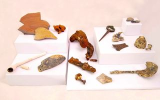 Foto van scherven, framgenten van metalen gebruiksvoorwerpen, een witte pijp met lange steel en andere voorwerpen in een vitrine.