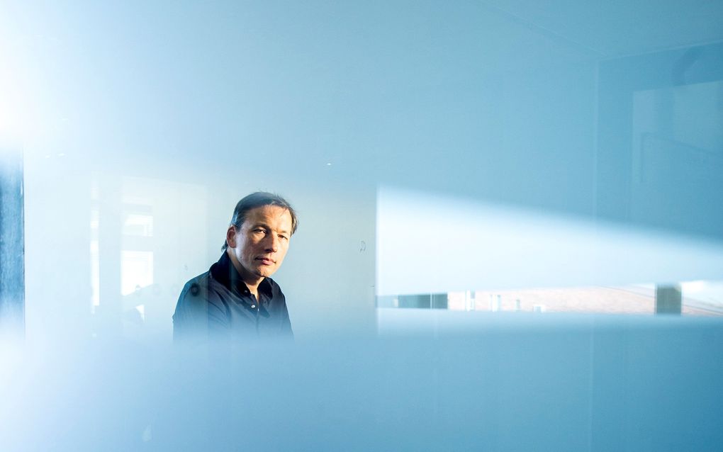 Man van middelbare leeftijd met donkerblauw overhemd zie je door een raam waar blinderende vlakken op zijn aangebracht; je ziet de man door een spleet tussen twee van die vlakken