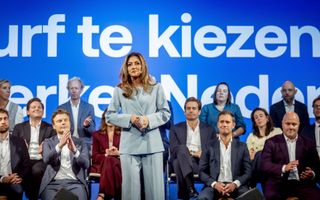 VVD-lijsttrekker Dilan Yeşilgöz in lichtgrijs pak houdt een toespraak tijdens een partijcongres van VVD. Achter haar zitten vooral mannen die voor haar applaudisseren. Op de muur staat Durf te kiezen