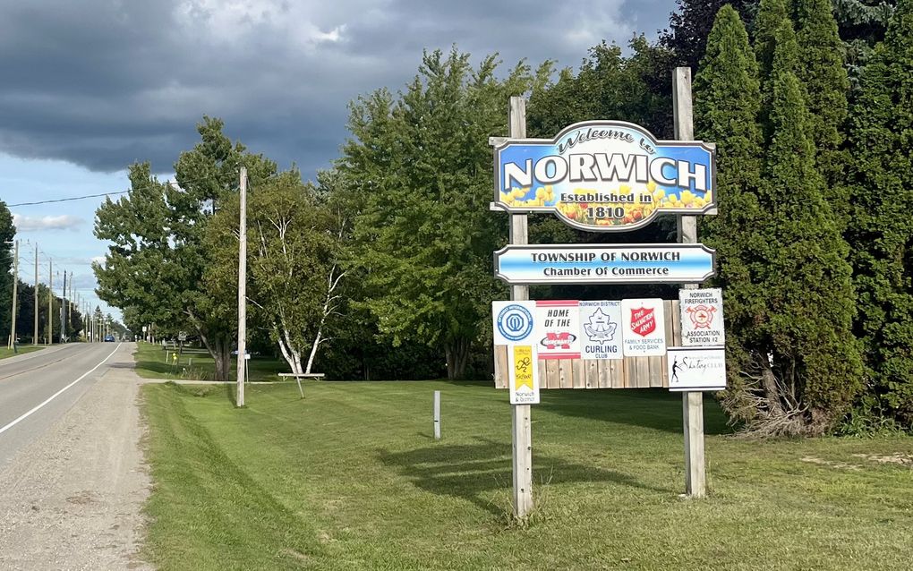 Een welkomstbord van een dorp in het gras naast een autoweg.