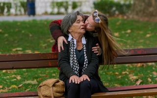 Een oudere vrouw zit op een bankje. Een jongere vrouw met zonnebril in haar lange blonde haren buigt zich vanachter het bankje over de oudere vrouw heen en geeft haar een zoen. Naast de oudere vrouw ligt een tas. 