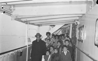 Zwart-witfoto van een groep donkere mensen met kinderen die over het gangboord van een schip lopen. Ze dragen tassen en zijn winters gekleed in dikke jassen en met hoeden en hoofddoeken. De foto is van voren genomen.