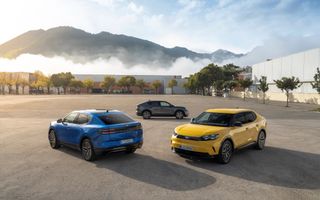 Een grijze, gele en blauwe auto staan op een grote vlakte. Langs de randen van het terrein staan grijzige loodsen, op de achtergrond wat bergen. Voor de loodsen staan groene bomen.