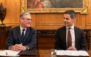 twee mannen in pak en das zitten achter een bruinhouten tafel en praten met elkaar 