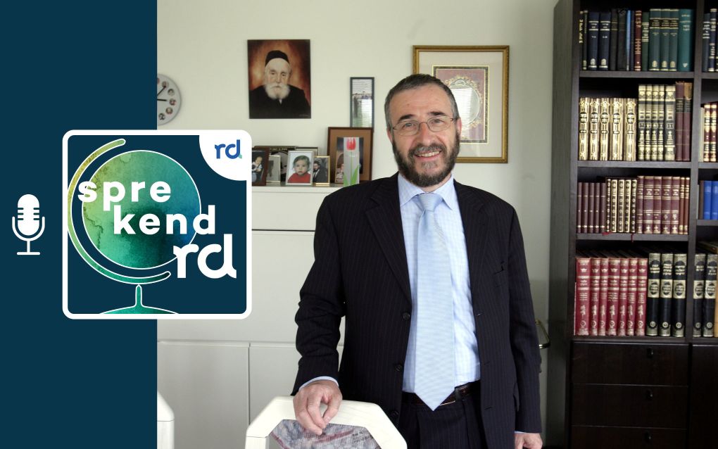 Man in pak voor een boekenkast en muur met schilderijtjes en foto’s.