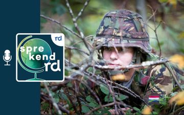 Een reservist in uniform zit laag bij de grond achter een aantal takken.