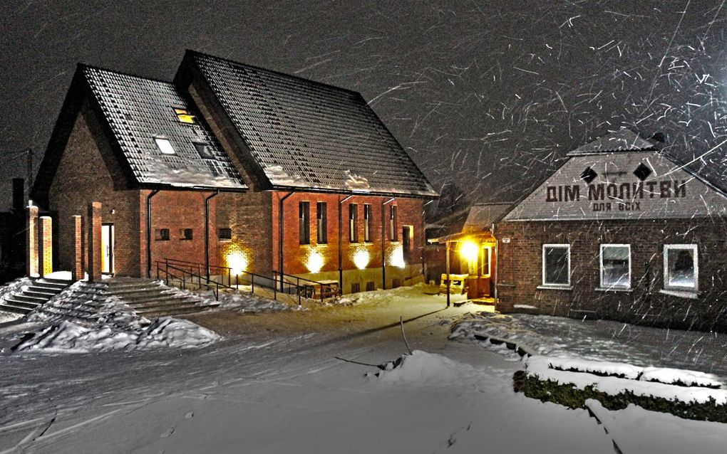 Verlicht kerkgebouw in de sneeuw. Ernaast staat een huis. 