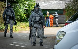 Zwaarbewapende politiemensen staan voor een boerenschuur. Op de achtergrond is een arrestant in een oranje overall te zien. 