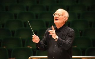 Oudere man met grijs haar heeft een baton in zijn hand en dirigeert een onzichtbaar orkest. Achter hem zijn lege groene stoelen te zien. 