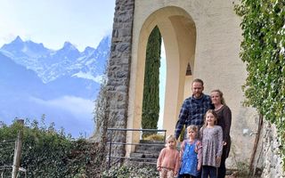 Jong gezin met drie meisjes staat voor een geel gebouw in een alpenlandschap. Links zijn hoge bergen te zien.