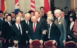 Groepsfoto van enkele tientallen politici. Op de voorgrond een vergadertafel met fraaie rode stoelen. Op de achtergrond enkele landenvlaggen.