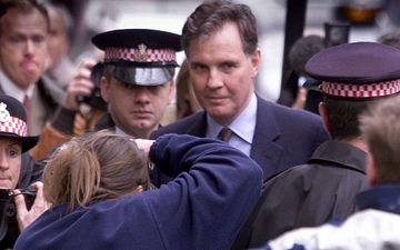 Een man in pak, te midden van verslaggevers en agenten, kijkt ongeveer richting de lens van de fotograaf.