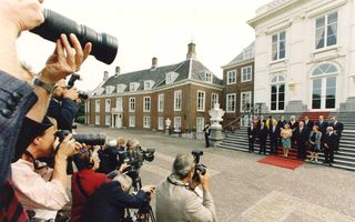 mannen in pak en vrouwen rond de koningin op de rode loper op de trap van het paleis. Vooraan zie je op de rug de fotografen met hun camera’s.
