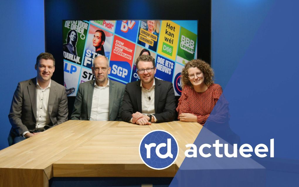 Drie mannen en een vrouw zitten aan een houten tafel. Achter hen is een scherm zichtbaar met verkiezingsposters.