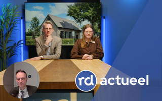 Een man en een vrouw zitten achter een houten tafel. Op de achtergrond is een foto van een kerkgebouw te zien.