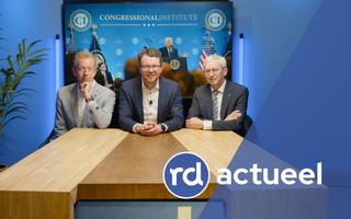 Drie heren in pak zitten vriendelijk glimlachend aan een houten tafel. Achter hen is een foto van de Amerikaanse president Donald Trump te zien.