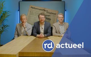 Drie heren, gekleed in een colbert, zitten achter een houten tafel. Achter hen is een afbeelding te zien van de eerste oplage van de krant uit 1971.