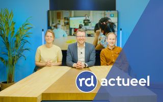 Drie redacteuren achter houten tafel in videostudio. 
