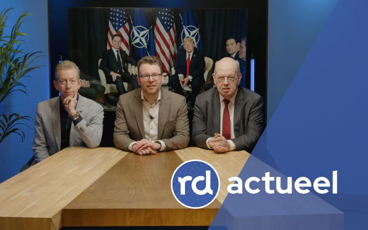 Drie mannen met een colbert aan zitten aan een houten tafel. Op de achtergrond is een scherm te zien met een foto van Rutte en Trump.