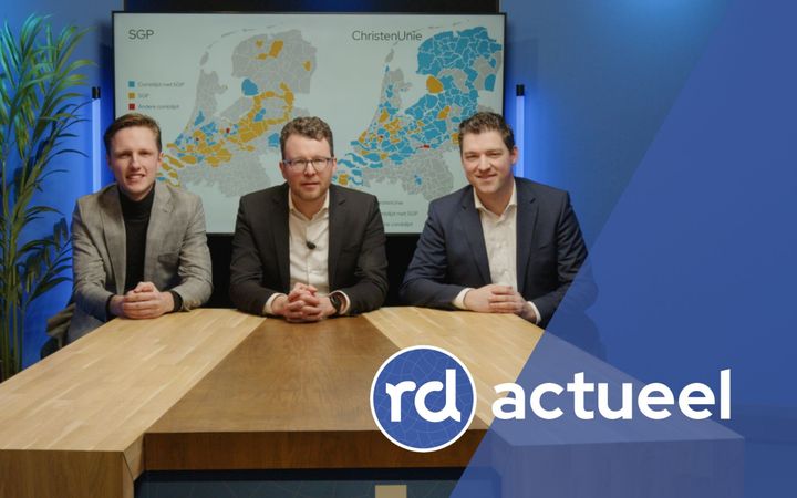 Drie vriendelijk lachende mannen met colberts aan zitten aan een houten tafel. Achter hen is een kaart te zien van Nederland.