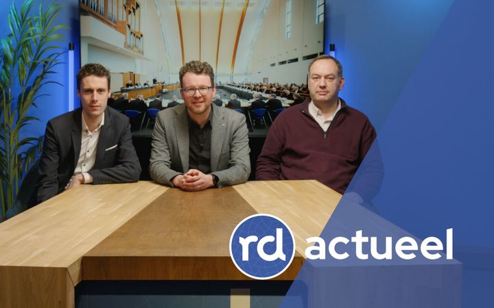 Drie vriendelijk ogende en net geklede heren zitten aan een houten tafel.