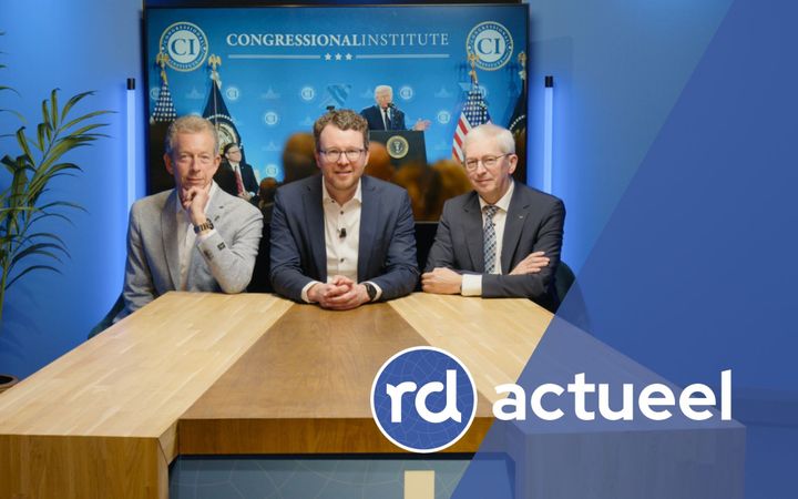 Drie heren in pak zitten vriendelijk glimlachend aan een houten tafel. Achter hen is een foto van de Amerikaanse president Donald Trump te zien.