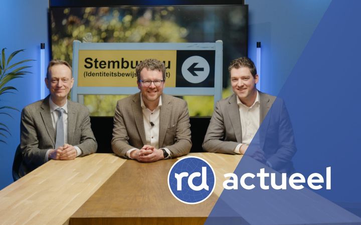 Drie vriendelijk lachende mannen zitten aan een houten tafel. Achter hen is een foto te zien van een bord met daarop de tekst: ‘Stembureau’.