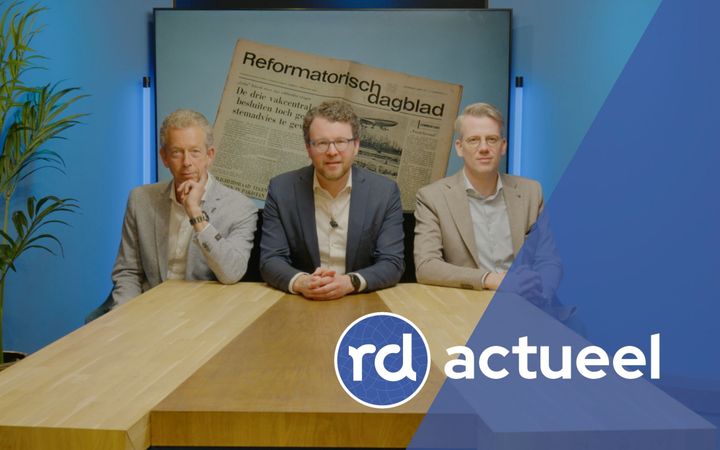 Drie heren, gekleed in een colbert, zitten achter een houten tafel. Achter hen is een afbeelding te zien van de eerste oplage van de krant uit 1971.