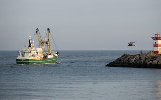Een groene viskotter vaart de zee op, langs een pier van grote betonblokken waar een rood-wit gestreept torentje met een havenlicht op staat.