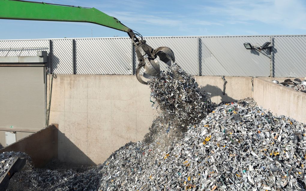 Een arm van een kraan met daaraan een grijper trekt een hoop oud metaal uit een hoop schroot. 