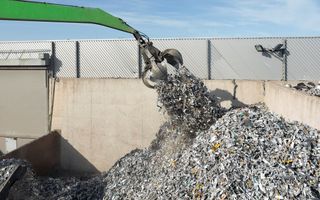 Een arm van een kraan met daaraan een grijper trekt een hoop oud metaal uit een hoop schroot. 