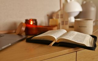 Een open Bijbel, een laptop en een koffiekopje staan op een lichtbruin kastje.