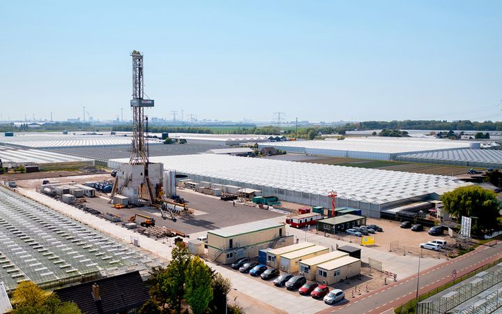 Een torenhoge installatie staat tussen tuinbouwkassen. Bij de installatie staan bouwketen en bedrijfsauto’s.