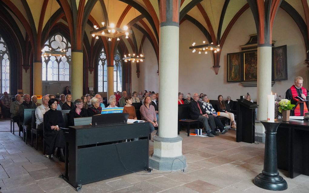 Kerkinterieur met mensen op stoelen en banken tussen pilaren. Vooraan staat een man in toga.