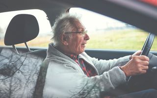 Oude man met grijs vest en rode geblokte blouse zit met een verwarde blik achter het stuur van een auto.