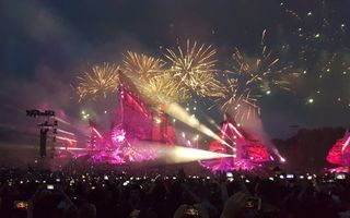 Een groot podium in de avond met lichten en vuurwerk waarvoor een menigte mensen staat met hun telefoon omhoog.