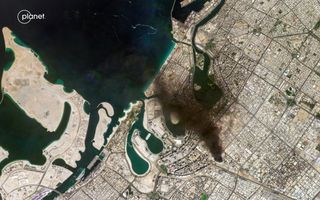 Satellietfoto waarop een zandkleurige stad en een groenblauwe zee te zien zijn. Boven de stad hangt een bruingrijze rookpluim