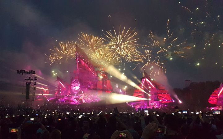Een groot podium in de avond met lichten en vuurwerk waarvoor een menigte mensen staat met hun telefoon omhoog.