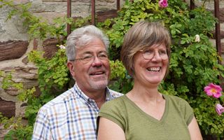 Man met grijs haar staat achter vrouw met bruin haar. Ze dragen beiden een bril. Ze staan voor een struik die tegen de muur van een huis groeit.