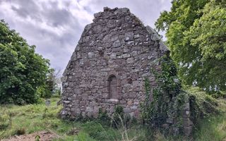 Op een grasveld tussen bomen staat de ruïne van een kerk. Er staat nog één muur overeind.