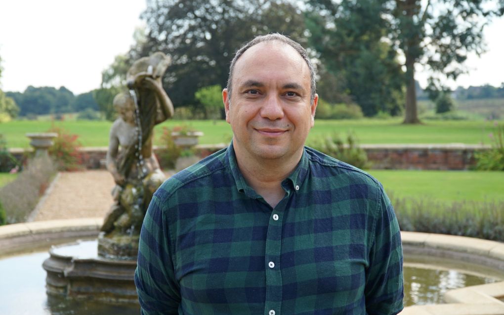 Portretfoto van een man met donker haar en een geblokte blouse. Hij staat in een park.