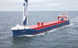 Een blauw vrachtschip met rode dekluiken vaart op zee. Voor op het schip staat een grote wit-blauwe vleugelvormige constructie.